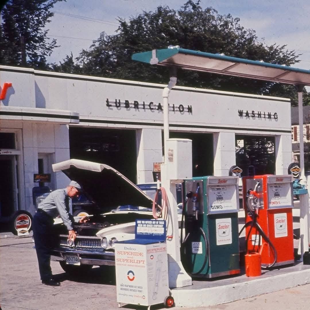 A man in uniform stands by a classic car with the hood open at an old-fashioned gas station with vintage pumps and a "Lubrication Washing" sign in the background.