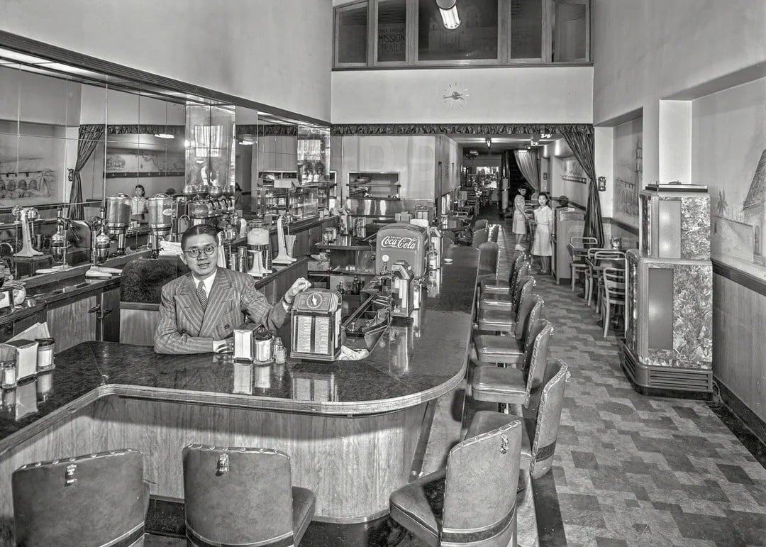 A man in a suit sits at the counter of a vintage diner with empty stools. In the background, staff in white uniforms stand near tables. The decor is classic, with checkered flooring and a visible Coca-Cola machine.
