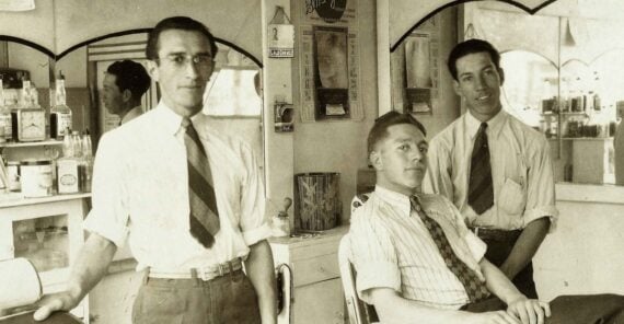 Three men in dress shirts and ties are in a vintage barbershop. One is seated in a barber chair while the other two stand nearby. The room has mirrors, shelves with products, and an old-fashioned atmosphere.
