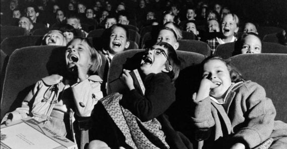Black and white photo of children sitting in theater seats, laughing heartily as they watch a movie. Pizza boxes and candy rest on their laps, conveying a joyful and carefree atmosphere.