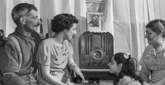 A vintage black-and-white photo of a family of four sitting together, listening to a radio. An older man, two women, and a young girl appear engaged and happy in a cozy, curtain-draped room.