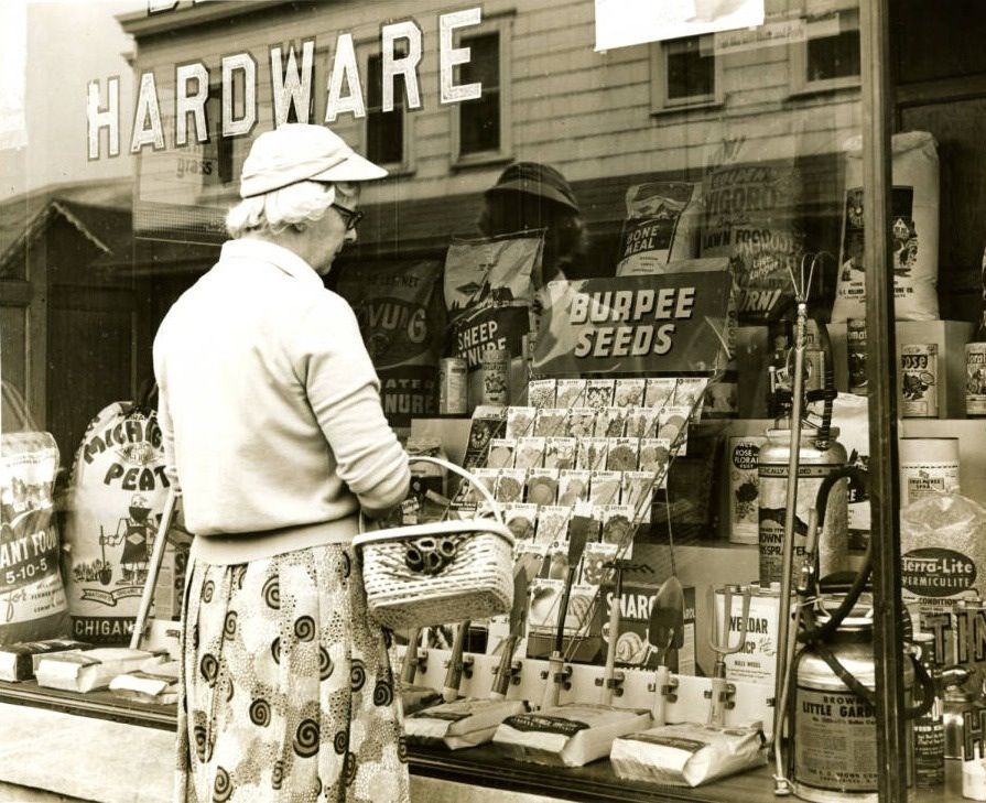 An elderly woman with a basket stands outside a hardware store window, looking at seed packets and gardening supplies displayed behind glass. The word "HARDWARE" is visible above the display.