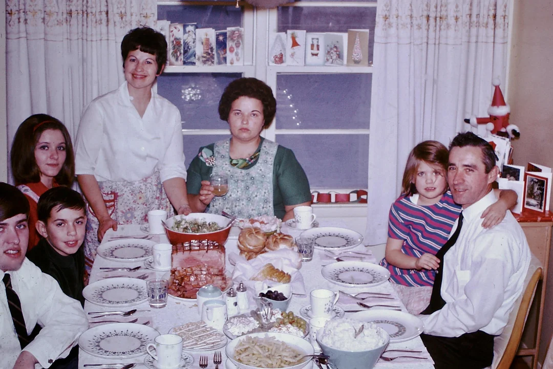 A family of seven sits and stands around a dining table set with food, plates, and mugs. Holiday cards and decorations are on the windowsill behind them. Everyone is smiling or looking at the camera.