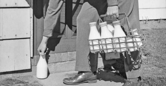 A person in dress shoes and trousers delivers glass bottles of milk, placing one on a doorstep while holding a metal carrier with more bottles, next to a building.