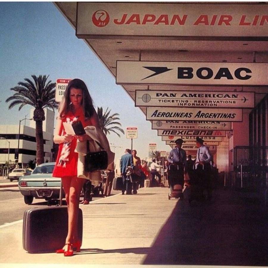 A woman in a red dress stands with a suitcase outside an airport terminal, reading a ticket. Overhead signs display airline names like JAPAN AIR LINES, BOAC, Pan American, and Mexicana. Palm trees and cars are in the background.