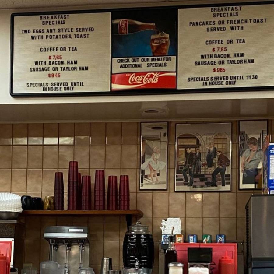 Menu board at a diner displaying breakfast specials, including eggs, potatoes, pancakes, bacon, ham, sausage, and toast. Coffee machines, cups, and kitchen items visible on the counter below.