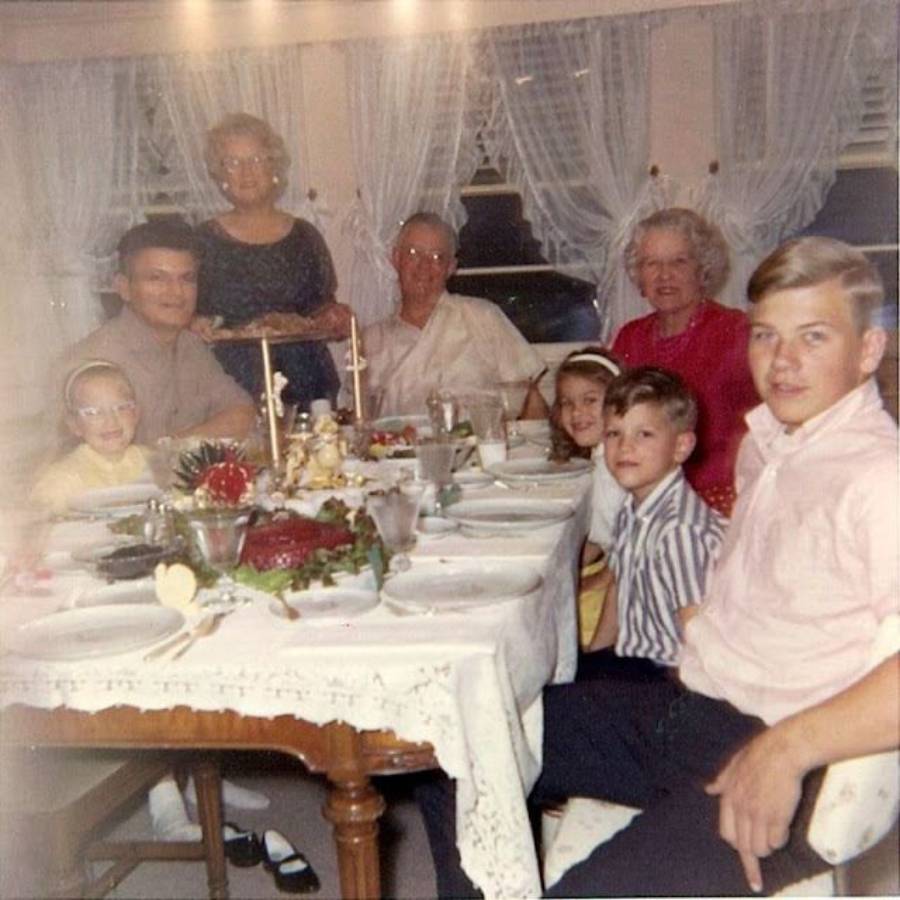 A family of eight, including kids and grandparents, sits around a dining table set for a meal in a cozy, curtain-lined room. Everyone is smiling and dressed in nice clothes.