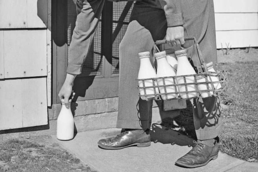 A person wearing dress shoes and holding a wire basket of glass milk bottles places one bottle on a doorstep outside a building.