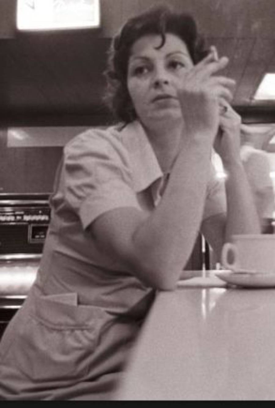 A woman in a diner uniform sits at a counter, holding a cigarette in one hand and looking away. A coffee cup is on the counter in front of her.