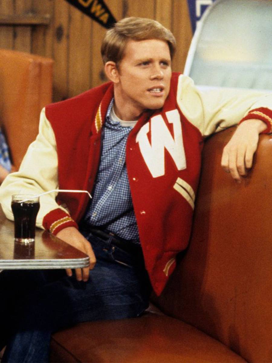A young man in a red and white letterman jacket sits in a diner booth with a drink and straw on the table, leaning back and looking to the side.