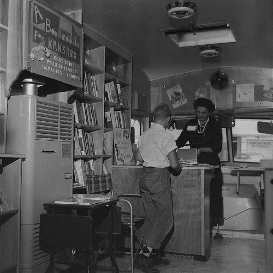 A boy checks out a book at a mobile library counter, staffed by a woman. Bookshelves line the walls, and a sign reads "A Bookmobile for Kansas." The setting appears to be inside a vehicle or trailer.