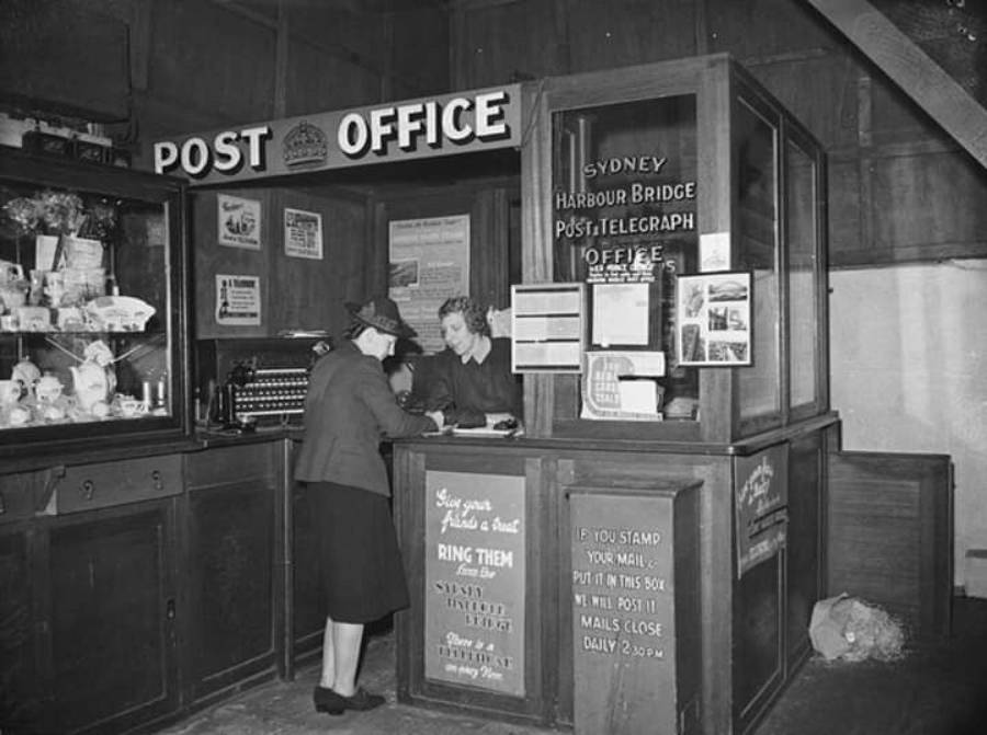 Black and white photo of two women at a vintage post office counter; signs mention Sydney Harbour Bridge Post/Telegraph, with postal items, notices, and a display case visible.