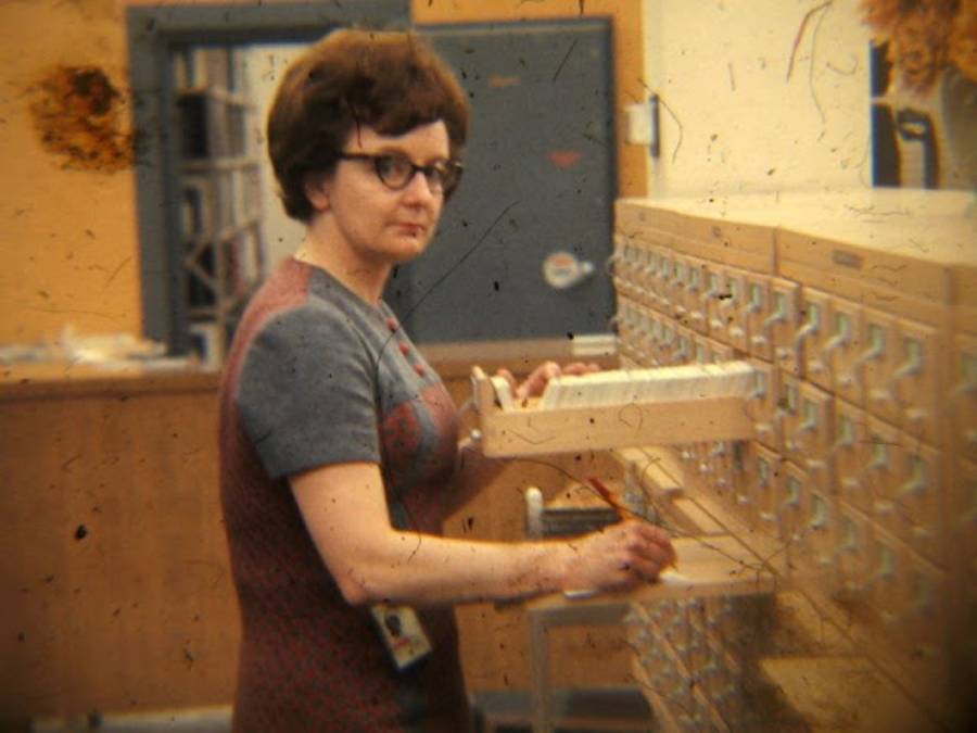 A woman wearing glasses and a patterned dress stands at a library card catalog, holding a card and a pencil, looking toward the camera. The setting appears to be a vintage or old-fashioned library.