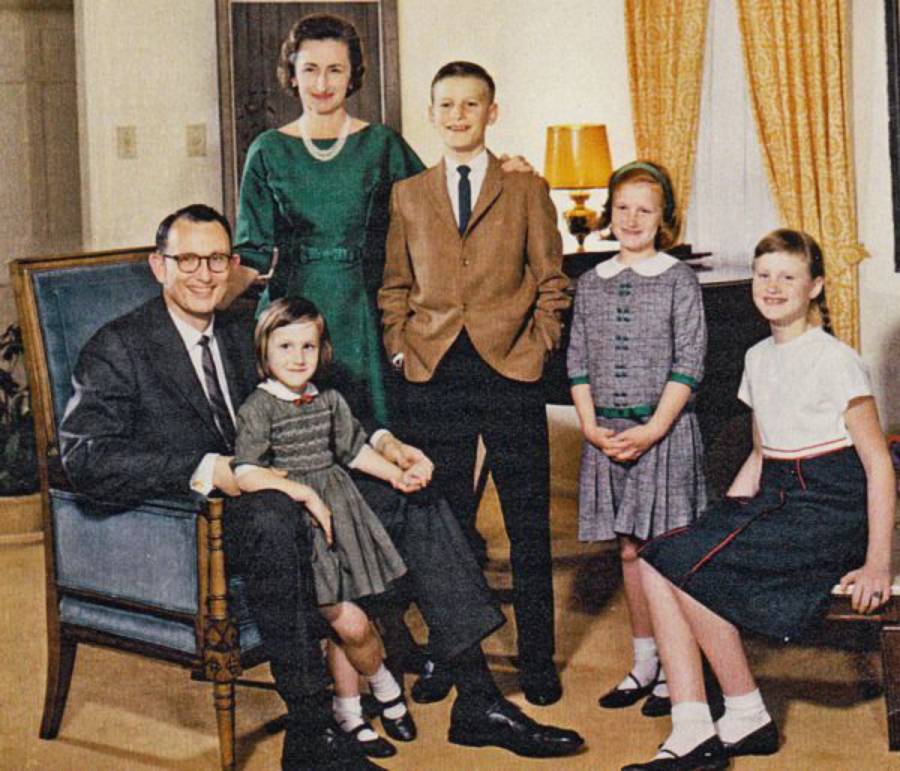 A family of six poses indoors; father seated with youngest daughter on his lap, mother standing behind, son and three daughters standing close by. Everyone is dressed formally, smiling, in a warmly lit living room.