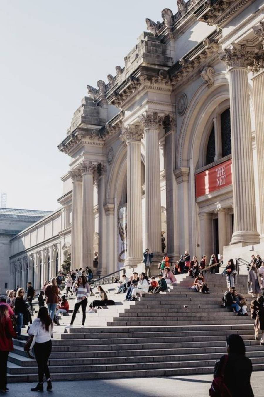 People sit and walk on the wide stone steps outside a grand building with tall columns and arched windows. The red sign reads "The Met," indicating it's the Metropolitan Museum of Art in New York City.