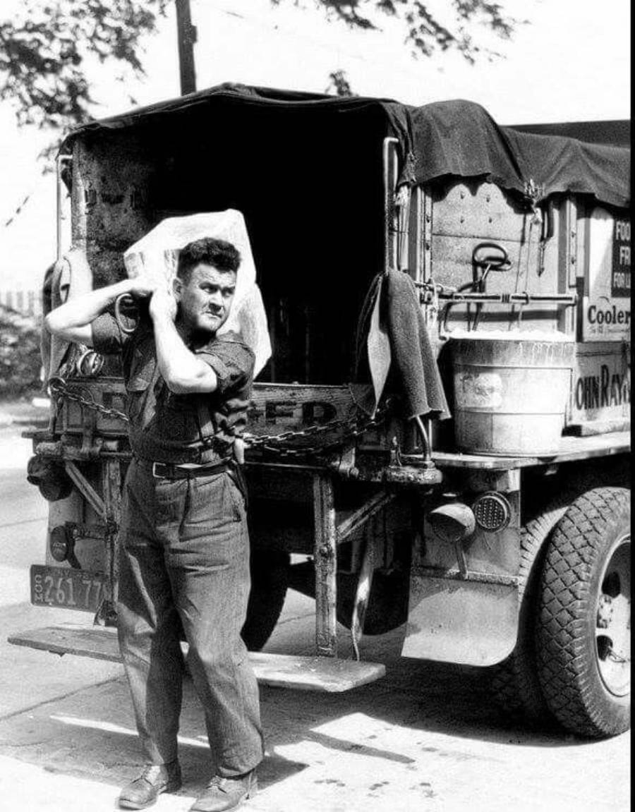 A man stands beside a truck, holding a large block of ice on his shoulder with both hands. The truck has an open back and various items inside, suggesting ice delivery. Trees and street signs are visible in the background.