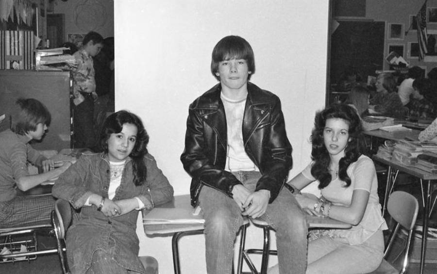 Three teenagers pose in a classroom; a boy in a leather jacket sits on a table between two girls. Other students are working at desks in the background. The image is in black and white.