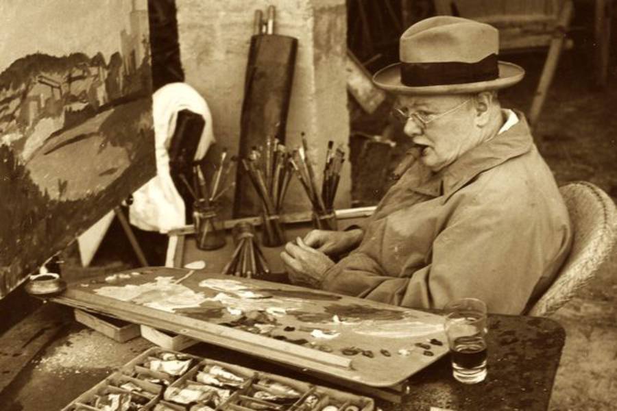 An older man in a hat and coat sits at an easel, painting outdoors. Art supplies, brushes, and a glass of water are on the table beside him. A landscape painting is visible on the easel.