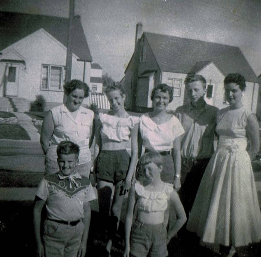 A group of five women and two boys pose outside in front of suburban houses on a sunny day, all wearing casual 1950s-style clothing and smiling at the camera.