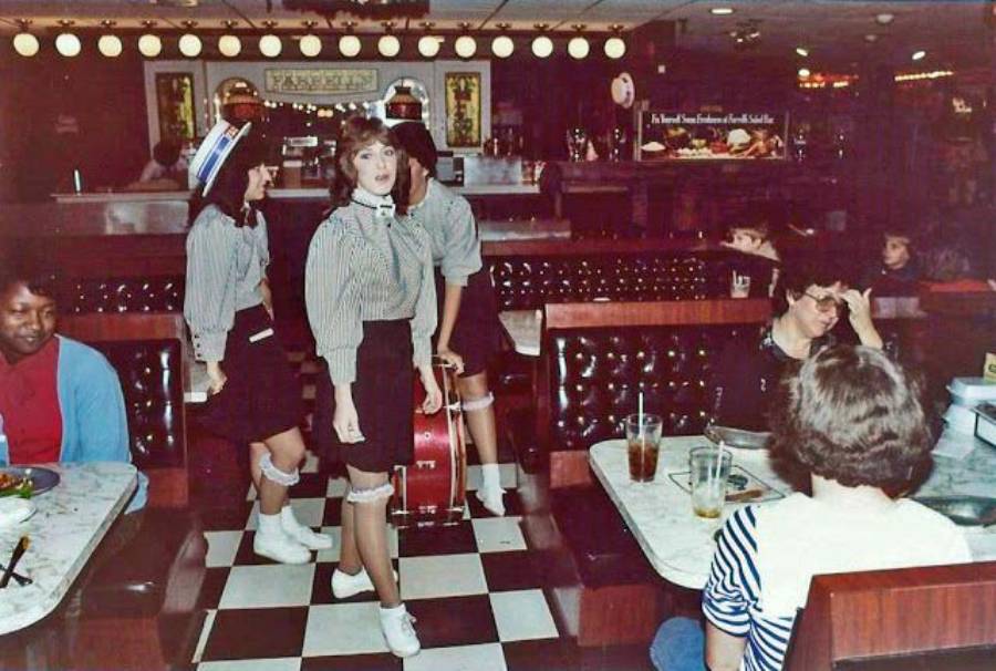 Three waitresses in vintage-style uniforms stand on a checkered floor in a busy retro diner with booths, drinks, and diners in the background.