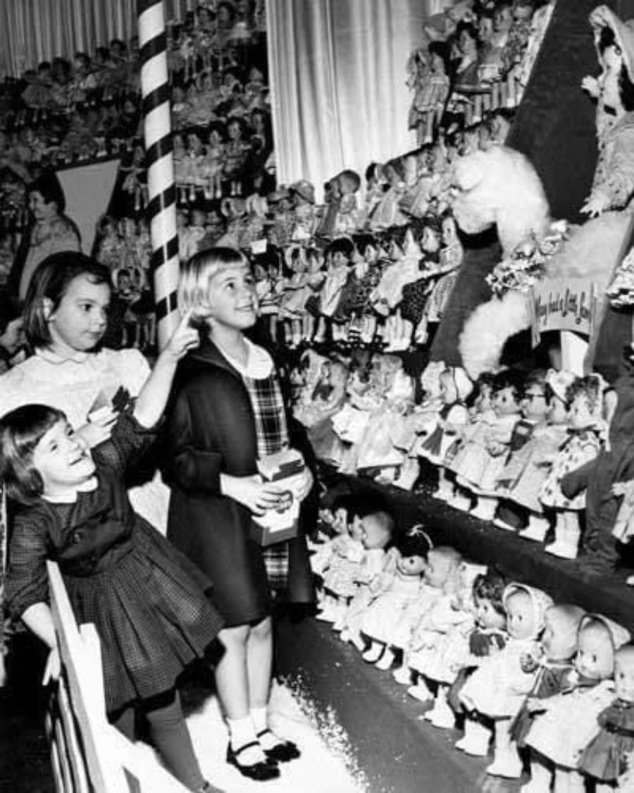Three children look excitedly at shelves filled with identical dolls in a store display. The scene appears festive, with decorative elements and dolls arranged in rows along the walls.