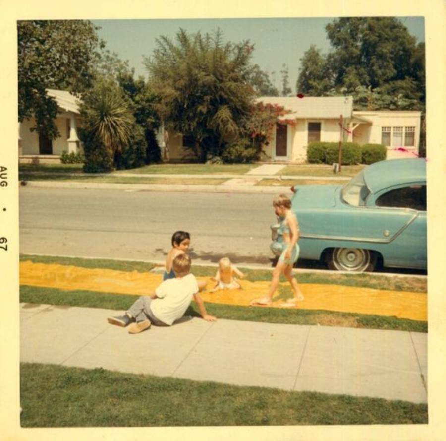 Four children play on a yellow slip-and-slide laid out on a suburban front lawn next to a sidewalk, with a blue vintage car parked nearby and houses and trees in the background. Photo is dated August 1967.