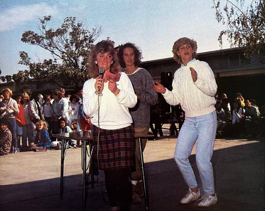 Three women stand in front of a table outdoors, one holding a microphone and singing, while the other two clap and dance. A crowd of people sits and watches in the background. Trees and a building are visible.