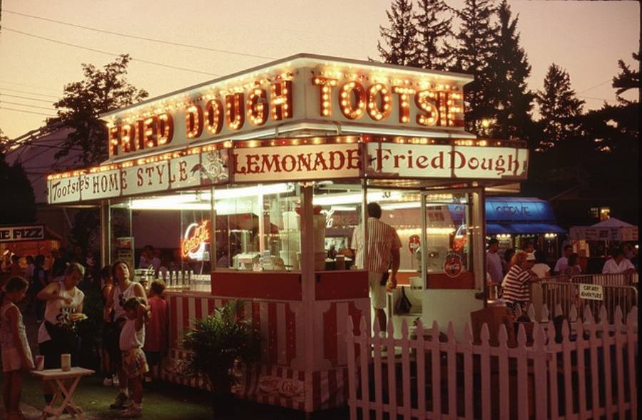 A brightly lit “Tootsie’s Fried Dough” food stand at dusk, with people gathered around, some ordering and others eating. The stand advertises lemonade and is surrounded by a white picket fence and trees in the background.