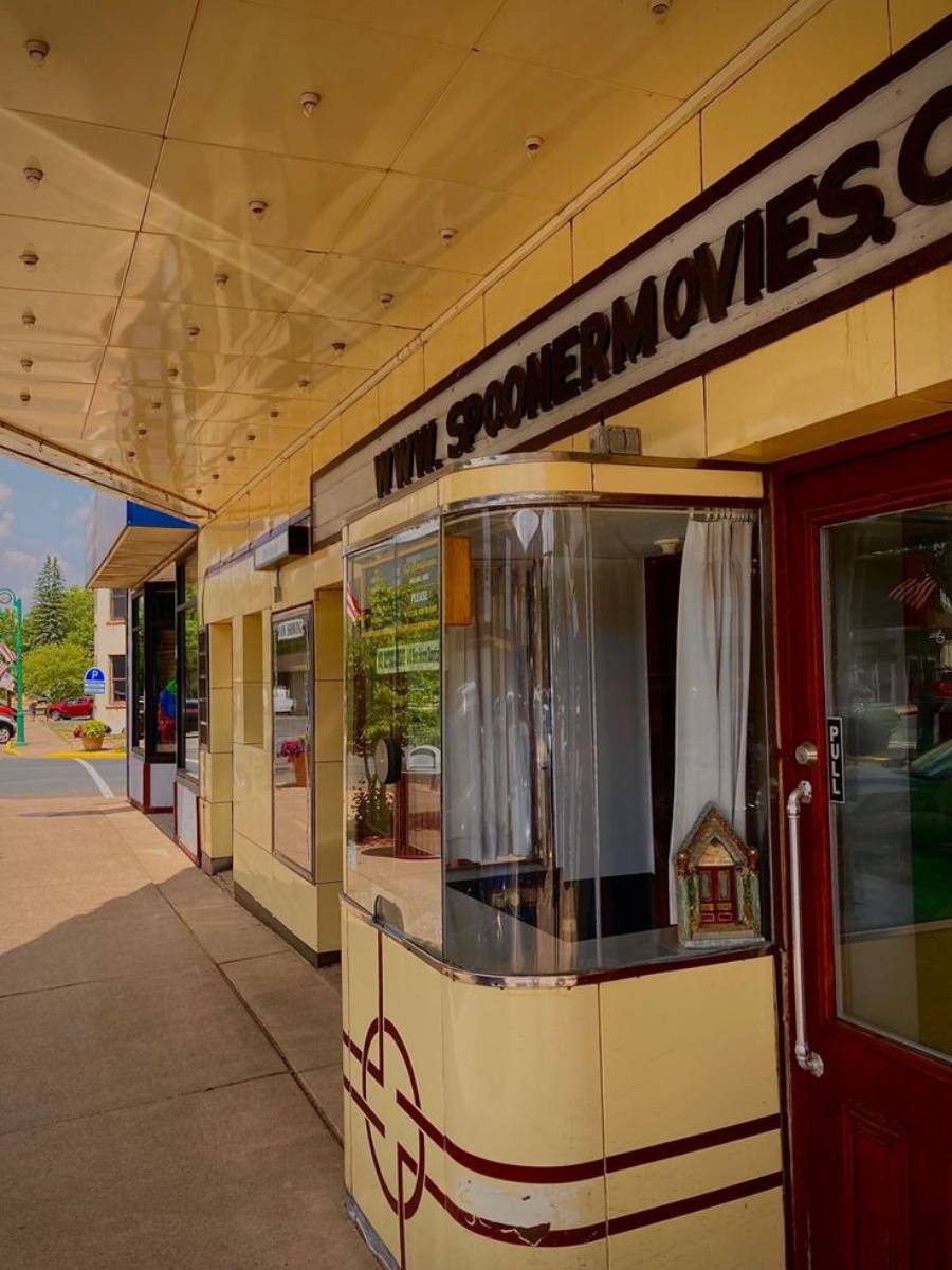 A vintage movie theater entrance with a yellow ticket booth, glass windows, and a marquee sign reading “SPOONER MOVIES.” The sidewalk and street are visible with trees and buildings in the background.