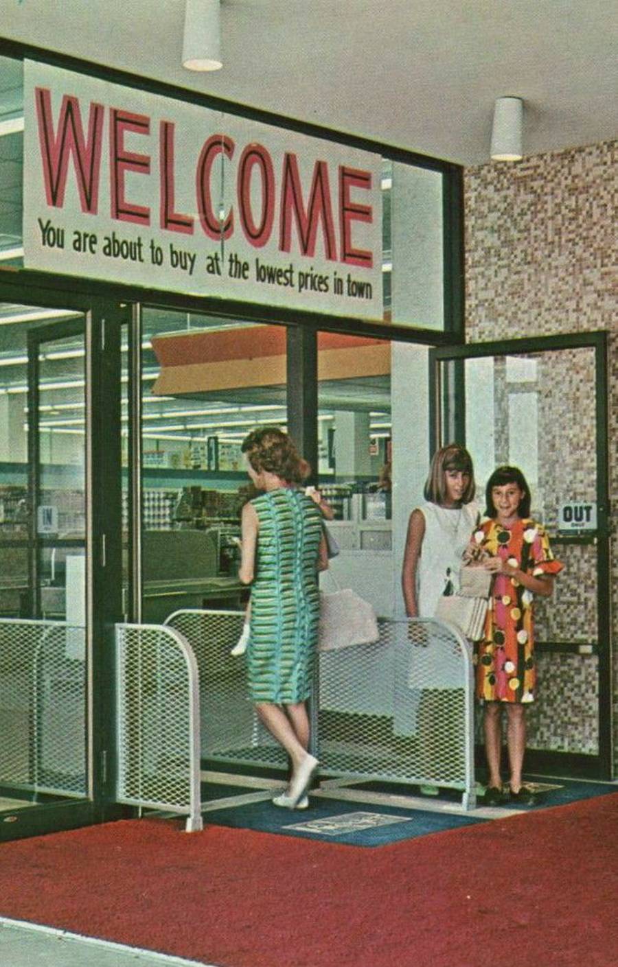 Three women exit a store under a large “WELCOME” sign that says, “You are about to buy at the lowest prices in town.” Two women stand talking; one walks away toward the door. The store interior appears bright and tidy.