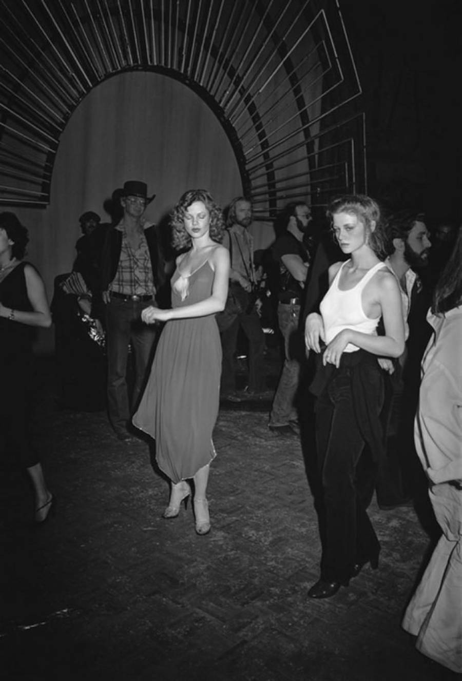 Black and white photo of two women in dresses standing on a dance floor, surrounded by people. An arched, decorative structure is behind them, with several onlookers in casual attire in the background.