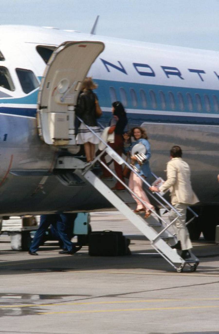 Several people are boarding a Northwest airplane via a stairway on the tarmac, with one person holding a newspaper and smiling women near the top of the stairs.