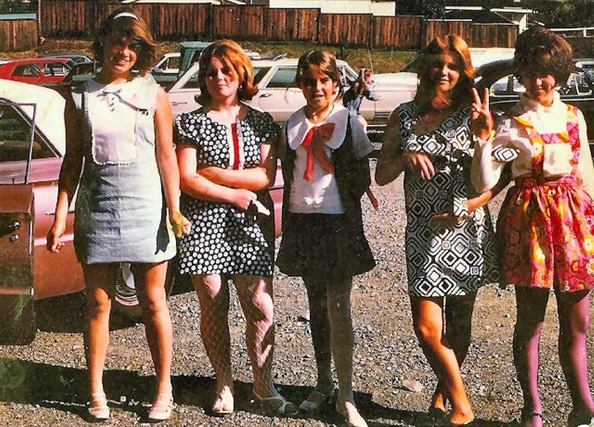 Five young women stand together outdoors in 1960s-style dresses, smiling and posing for the camera. There are cars and a wooden fence in the background, and the ground is gravel.