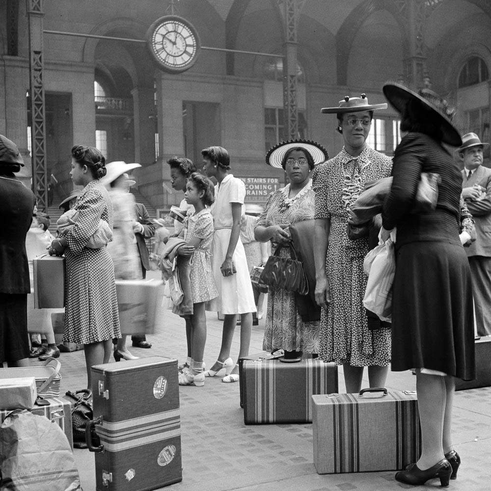 Black and white photo of women and children in dresses and hats, standing in line with suitcases at a busy train station. A large clock hangs above them.