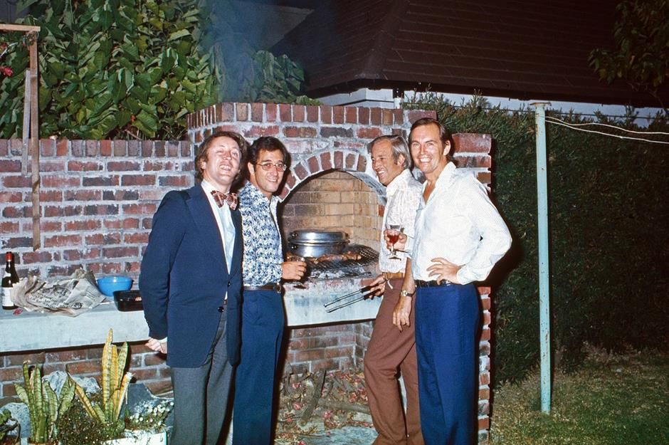 Four men stand smiling by a brick barbecue grill outdoors at night, dressed in 1970s-style clothing. They appear to be enjoying a casual gathering, holding drinks and grilling food in a backyard setting.