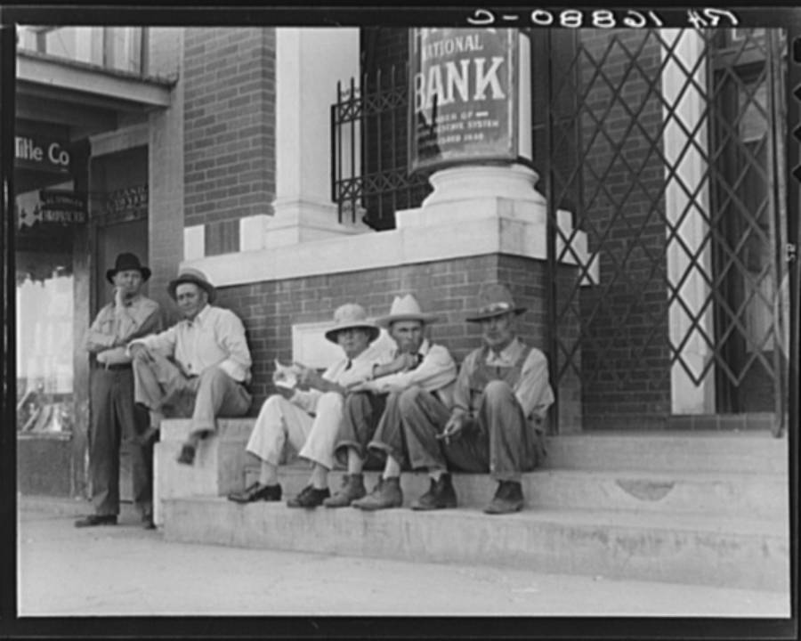 Five men in hats sit and lean on the steps of a brick building labeled "National Bank" in the background. They appear relaxed, possibly taking a break, on a sunny day.