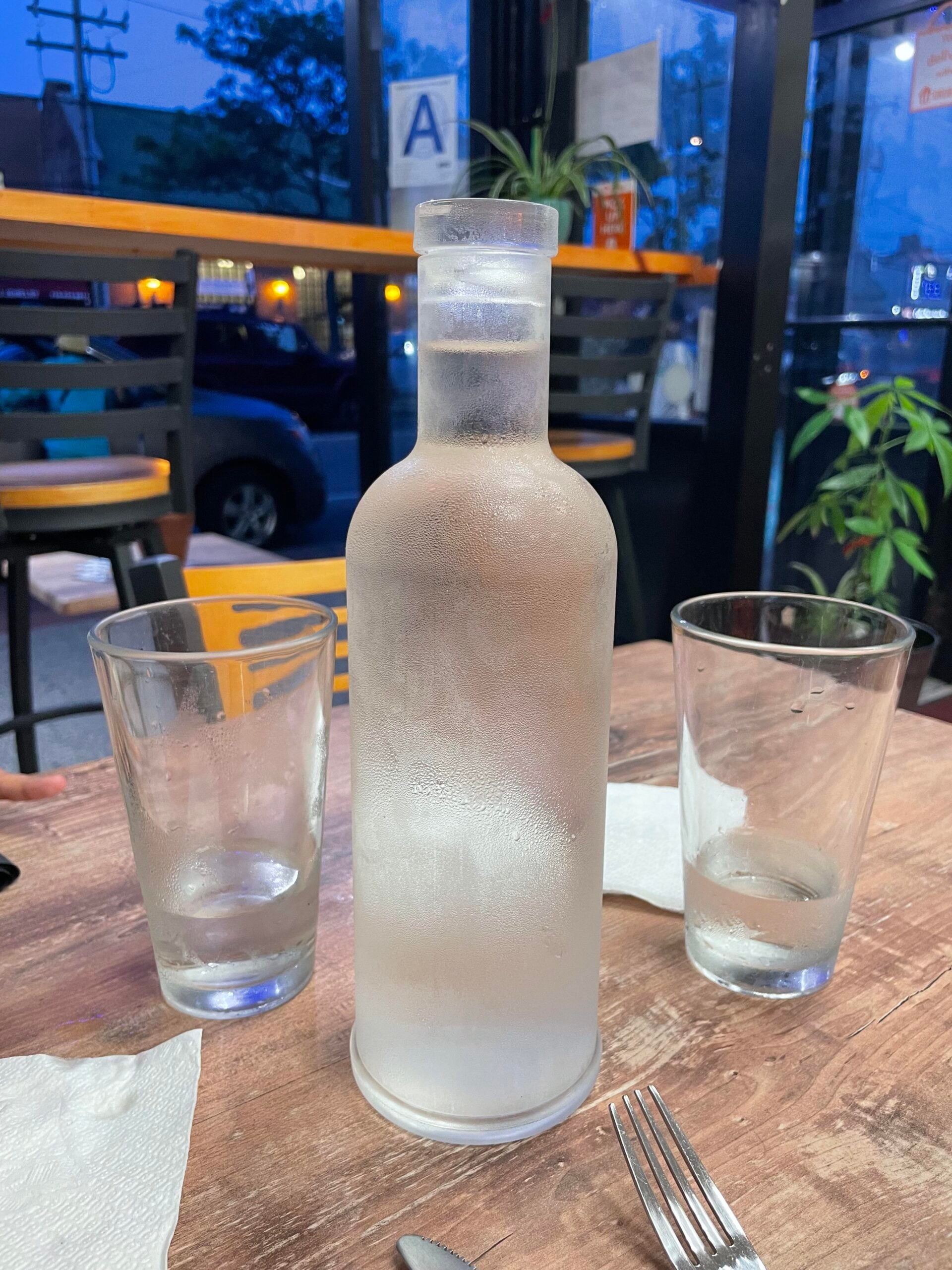 A frosted glass bottle of water and two half-filled glasses sit on a wooden table in a restaurant with chairs, napkins, a fork, and nighttime city view outside the window.