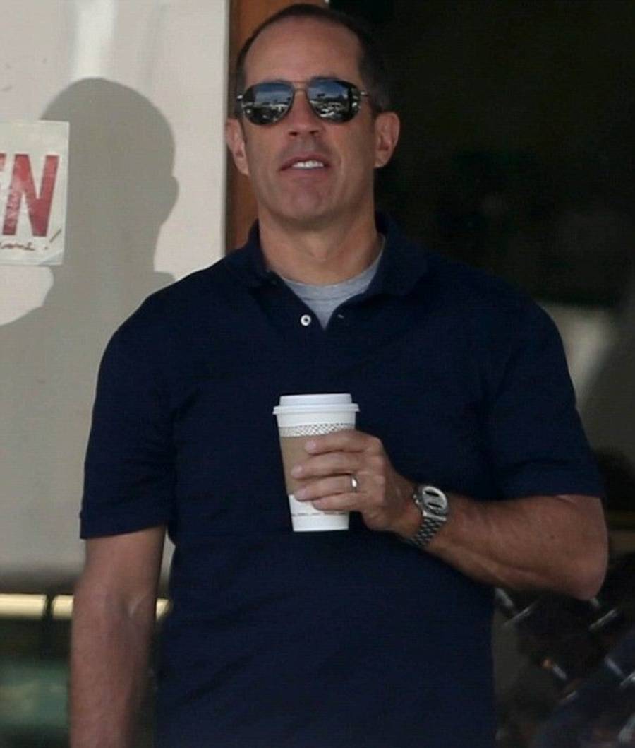 A man wearing sunglasses and a dark polo shirt stands outside, holding a coffee cup in front of him with one hand. He appears relaxed and is looking forward.