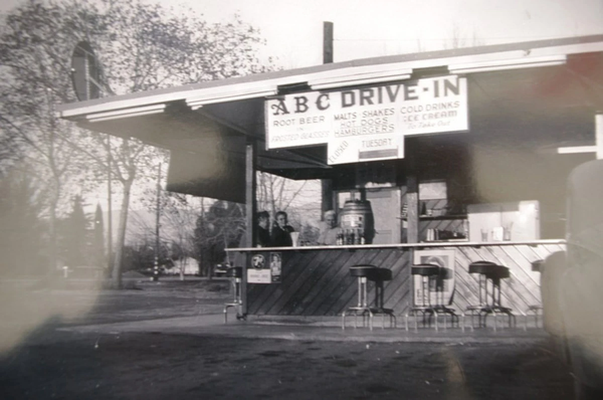 Black-and-white photo of a small, open-air drive-in stand called "ABC Drive-In," with stools, signs for root beer, malts, hot dogs, hamburgers, and ice cream. Two people stand behind the counter. Trees surround the area.