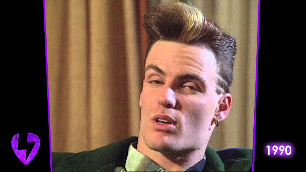 A person with a tall, styled haircut, wearing an earring and dark clothing, sits in front of beige curtains. The year "1990" appears in neon text in the lower right corner.