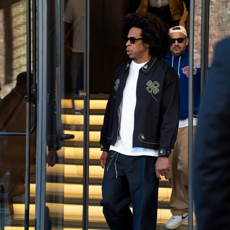 A man with curly hair wearing sunglasses, a black jacket with green embroidery, a white shirt, and dark pants exits a building with glass doors and illuminated steps. Another man in a hoodie and cap follows behind him.