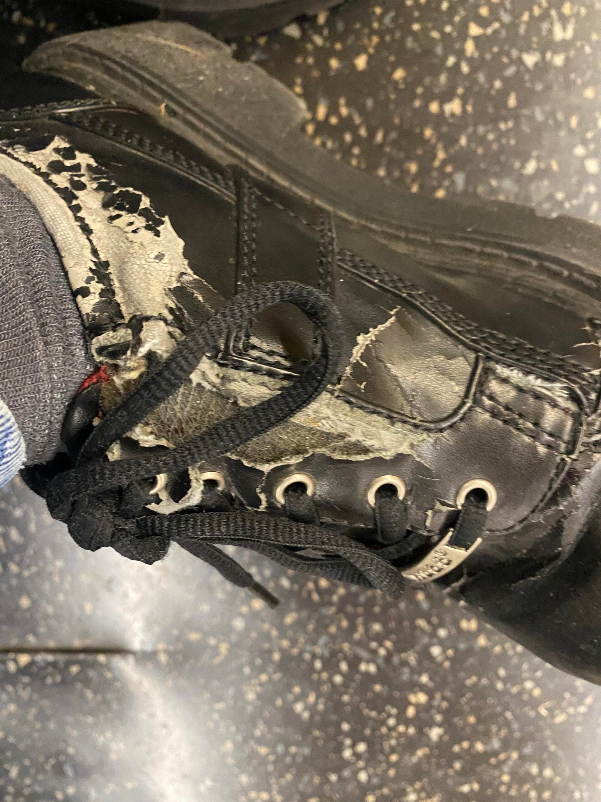 A close-up of a worn black sneaker with peeling outer material and frayed edges, showing visible damage. The shoe is being worn with a gray sock. The background appears to be a speckled floor.