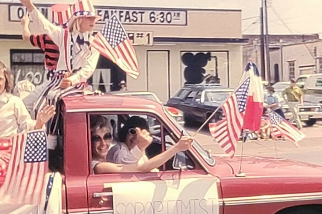 A parade scene shows people in a red truck holding American flags; one person is dressed in patriotic attire and waving. The background includes a diner with a "Breakfast 6:30 AM" sign.