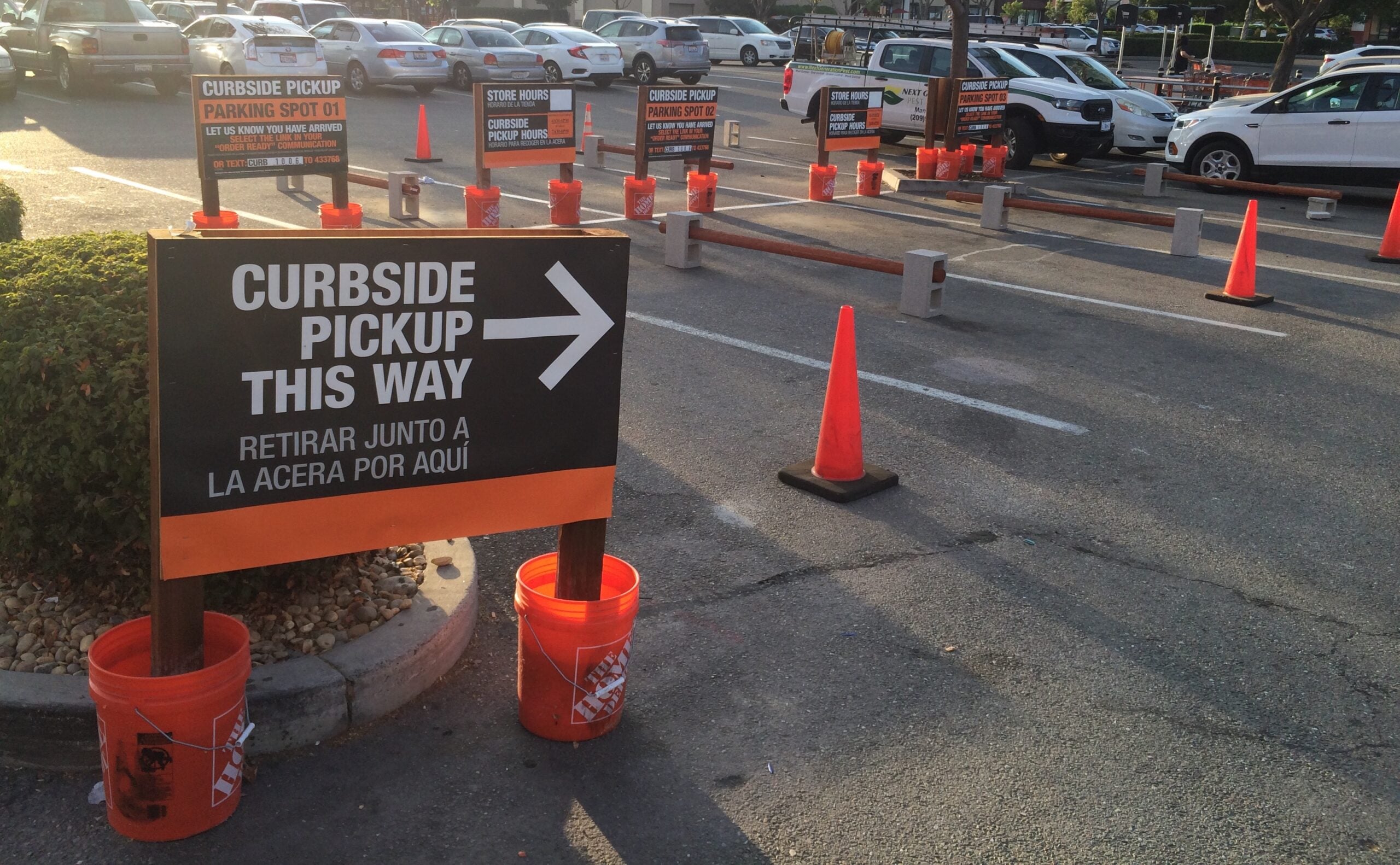 Parking lot with orange cones and signs directing drivers to curbside pickup spots. The largest sign reads "Curbside pickup this way" with a right arrow and Spanish text below. Several cars are parked in the background.