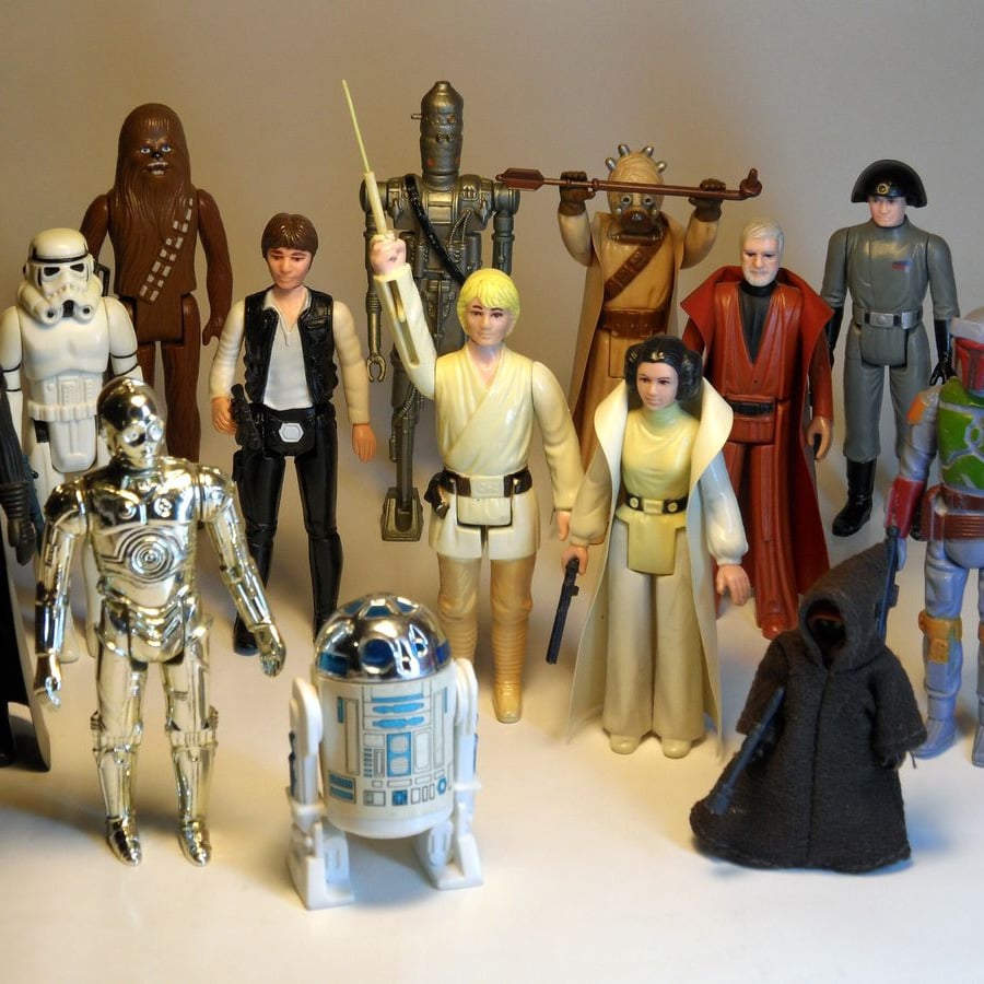 A group of vintage Star Wars action figures, including characters like Luke Skywalker, Princess Leia, Darth Vader, R2-D2, and more, arranged standing together against a plain background.