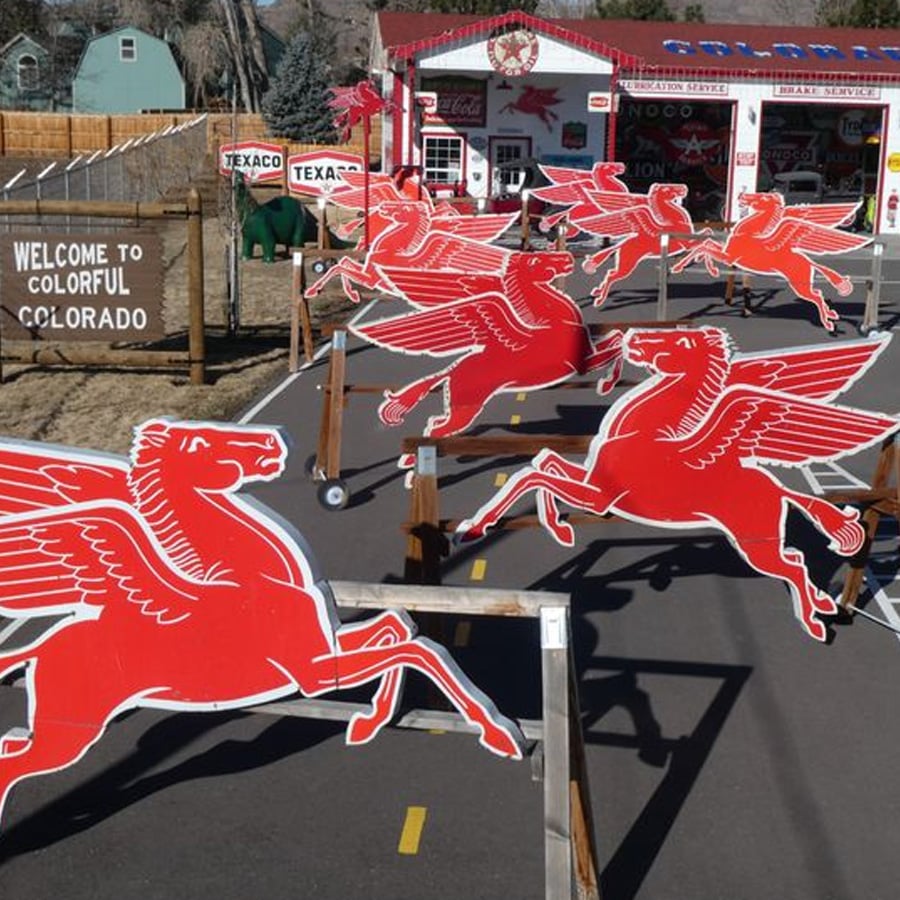 Several large red Pegasus signs are lined up on a mini road in front of a vintage-style gas station with a "Welcome to Colorful Colorado" sign visible on the left.