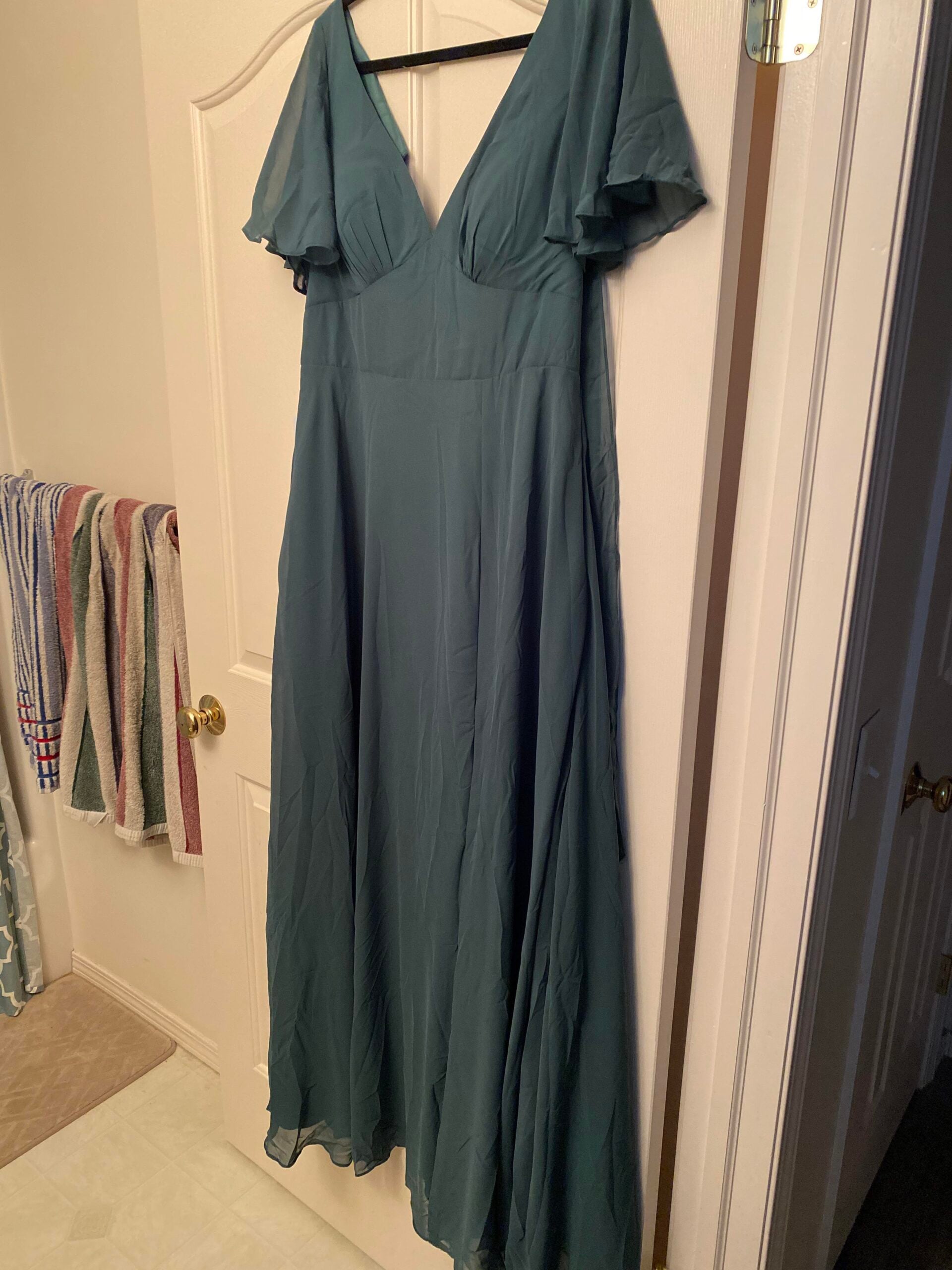 A long, teal green dress with short flutter sleeves and a V-neckline hangs on a white door in a bathroom. Towels are visible on a rack to the left.