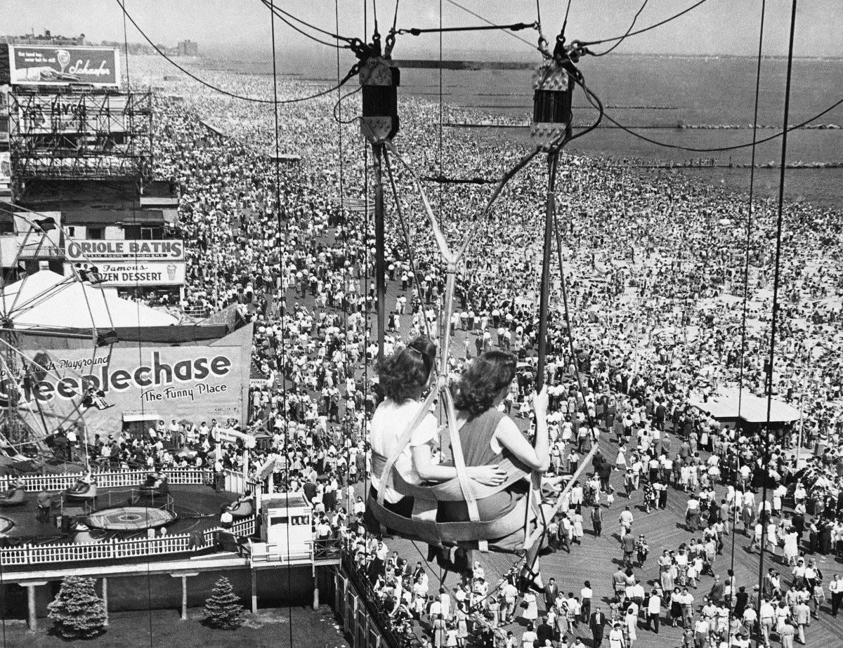 Two women ride a swing ride above a massive, crowded beach and boardwalk, with signs reading "Oriole Baths" and "Steeplechase" visible among the sea of people near the ocean.