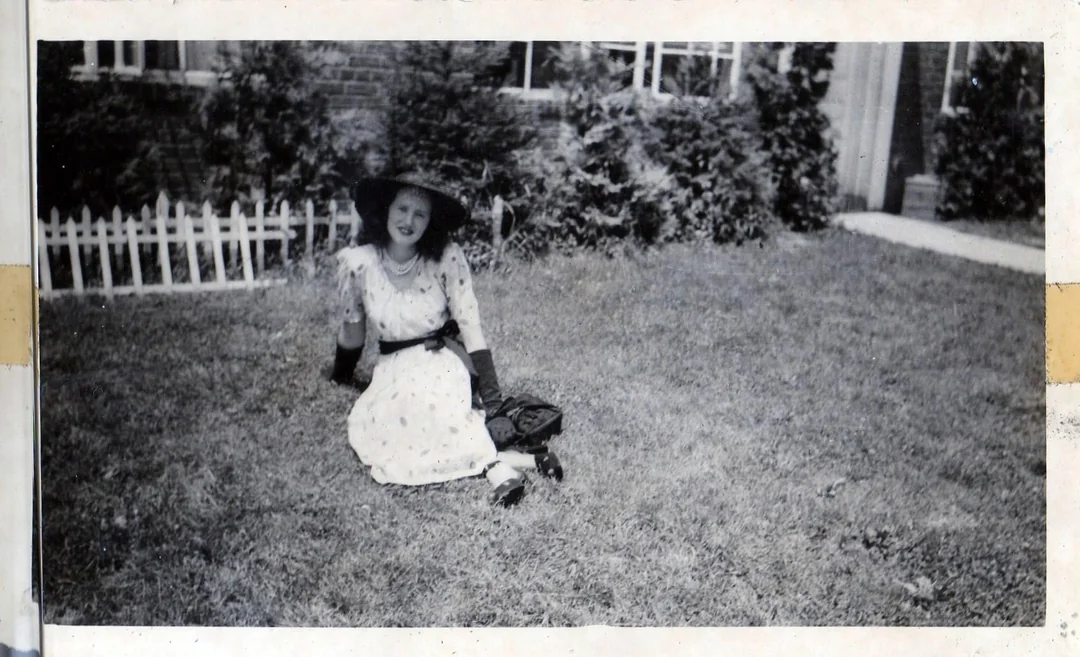 A woman in a wide-brimmed hat and dress sits on grass, smiling and waving, with bushes, a white picket fence, and a brick building in the background.