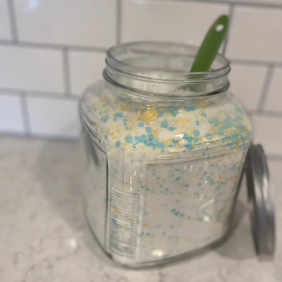 A glass jar filled with white powder mixed with blue and yellow beads sits on a counter. Its metal lid rests beside it, and a green plastic scoop is inside the jar. White tile backsplash is visible in the background.